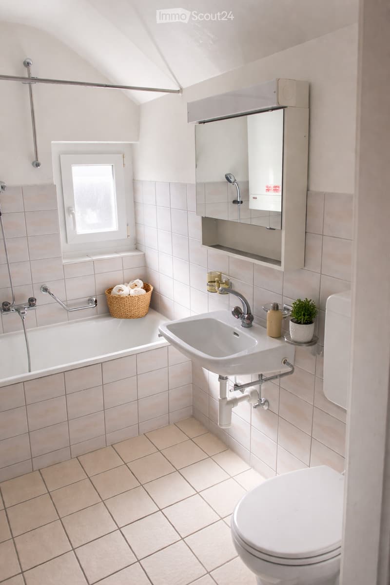 Bright bathroom with white tiles, a bathtub, sink, toilet, mirror cabinet, and a small window letting in natural light.