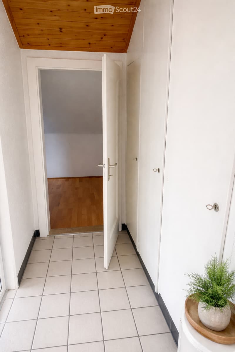 A narrow hallway with white tiled flooring and white walls, featuring a wooden ceiling and an open door leading to another room with wooden flooring.