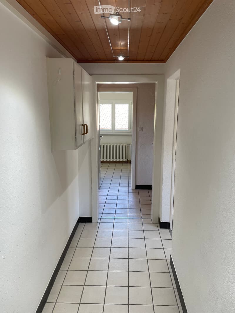 Empty hallway with tiled floor, white walls, and a ceiling light fixture. A door leads to another room with a radiator and window.