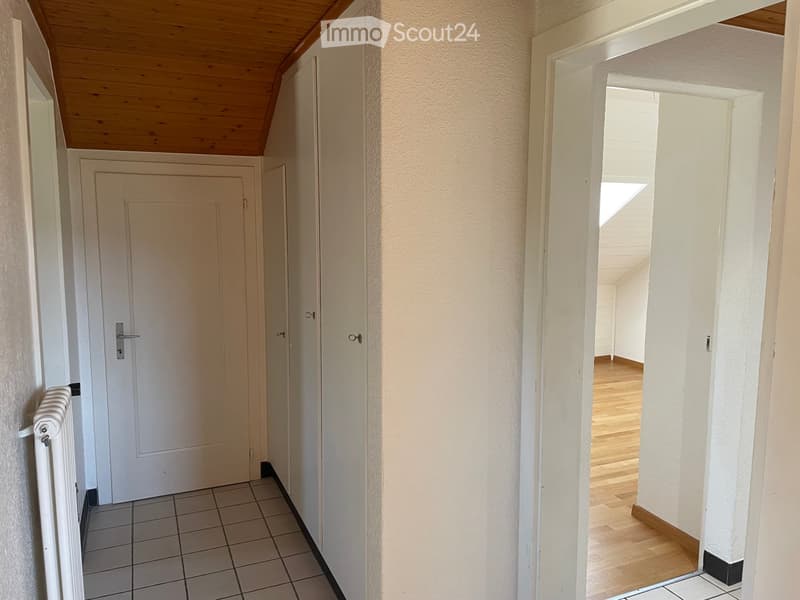 A narrow hallway with white walls, tiled floor, and wooden ceiling. Features a white door on the left and an open doorway leading to a room with wooden flooring on the right.