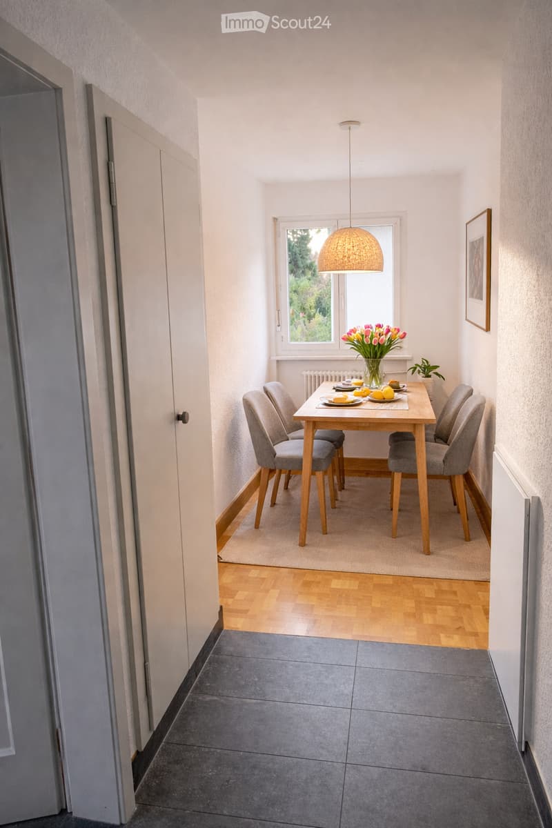 A small, well-lit dining area with a wooden dining table, four grey chairs, a vase with tulips, and a picture frame on the wall. The room features a mix of wooden and tiled flooring.