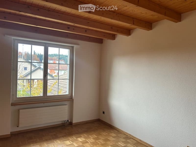 Empty room with wooden floor, white walls, large window with view of houses, radiator below window.