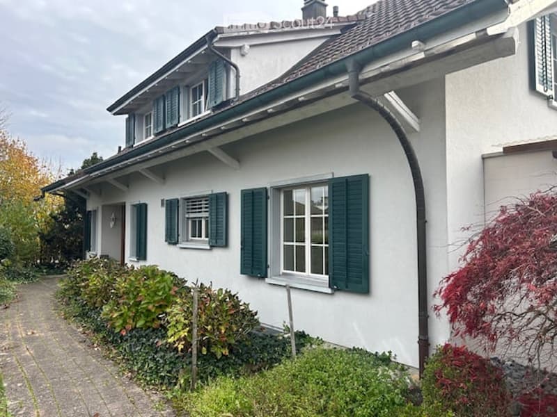 Two-story house with white exterior, green shutters, and multiple windows. The house has a sloped roof and is surrounded by a well-maintained garden with shrubs and trees.
