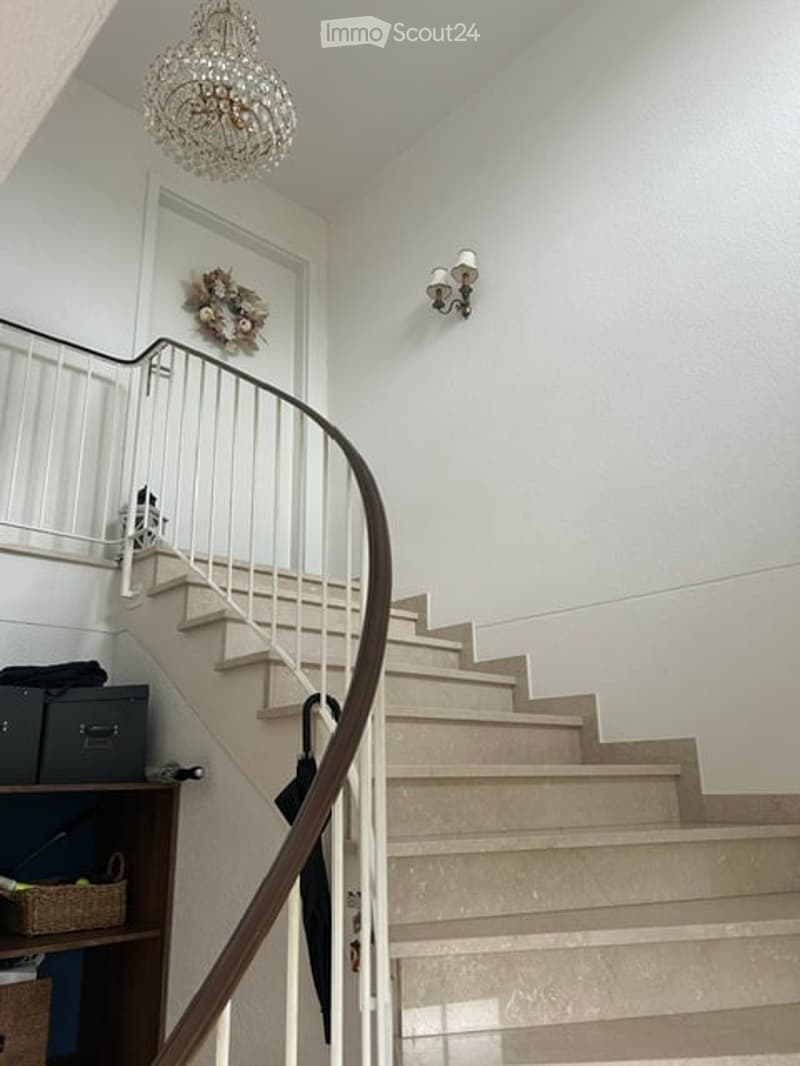 Spiral staircase with white railings and beige steps, chandelier hanging from the ceiling, wall-mounted sconce light, small shelf with items, umbrella leaning against the wall.