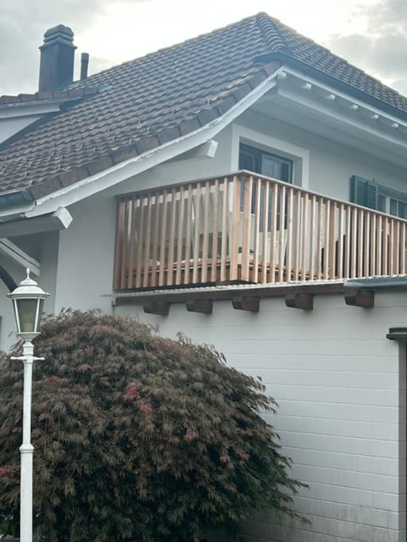 Two-story house with a balcony featuring wooden railings and a small table and chairs. The house has white siding and a tiled roof.