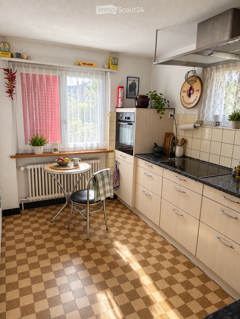 The kitchen features light wooden cabinets, a black countertop, and a checkered brown tile floor. A small round table with a black chair is placed near the window, which has sheer white curtains. The kitchen includes an oven, a refrigerator, and various s