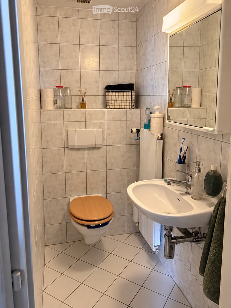 Modern bathroom with beige tiled walls and floor, featuring a toilet with a wooden seat, a sink with a chrome faucet, a mirror with built-in lighting, and various toiletries neatly arranged on shelves and the sink counter.