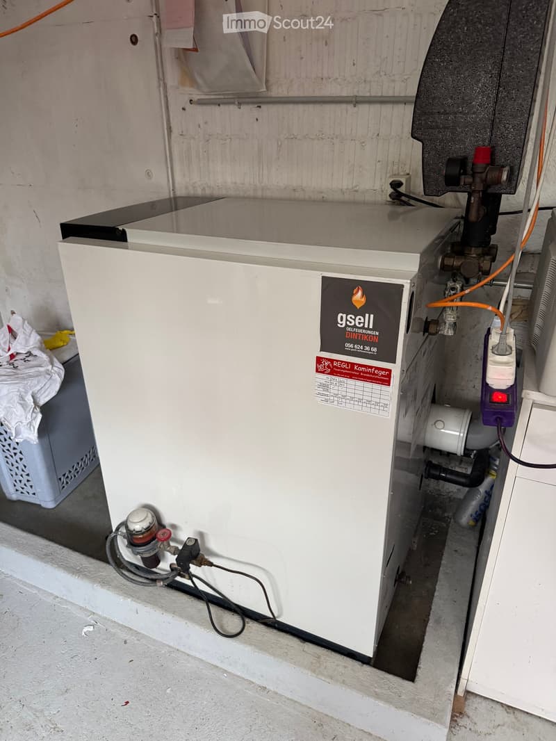 A white oil boiler with visible pipes and valves, installed in a utility space with a concrete floor and exposed wall.