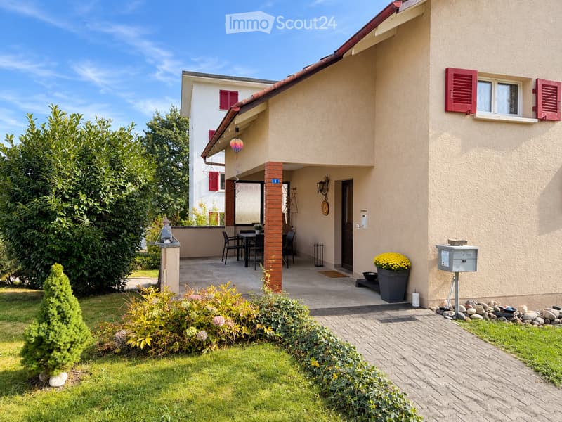 Two-story house with beige exterior, red shutters, and a covered patio area with a dining set, surrounded by a well-maintained garden and lawn.