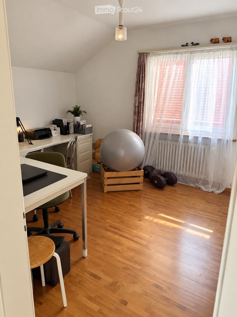 A well-lit room with wooden flooring, a large window with curtains, a desk with a chair, a ball on a wooden crate, and a small plant on the windowsill.