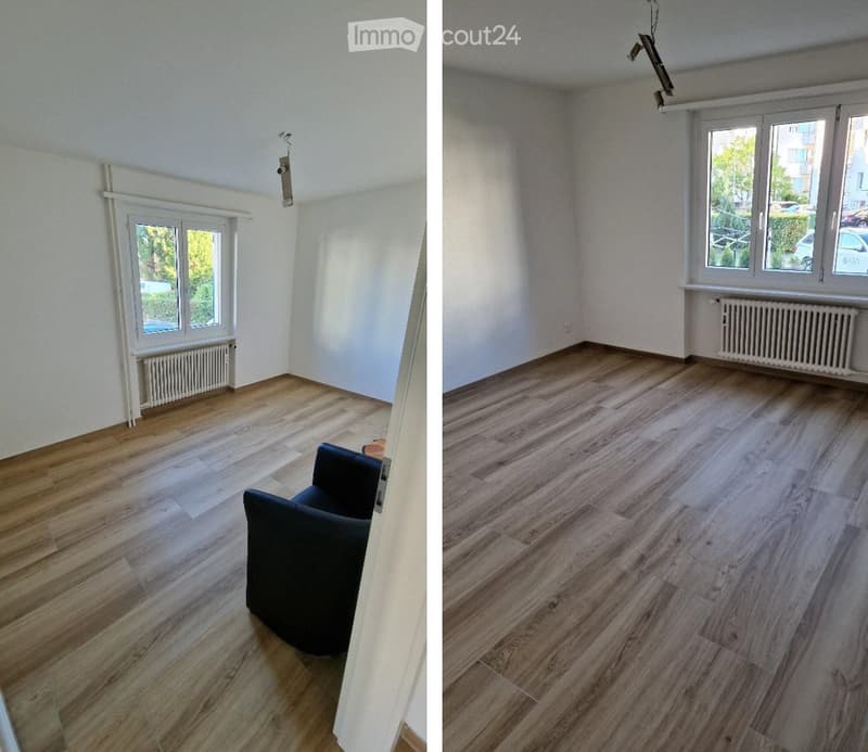 The room features wooden flooring, white walls, and large windows with views of greenery. A black sofa is positioned near the window, and a radiator is visible below the window on the left side.
