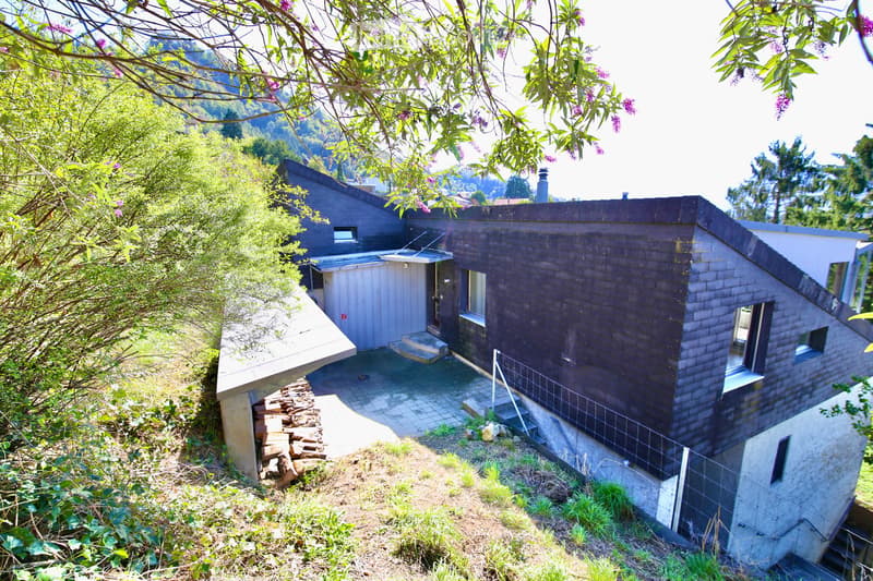 Modern, multi-level house with dark wooden exterior, situated on a sloped terrain with surrounding greenery and a small patio area.