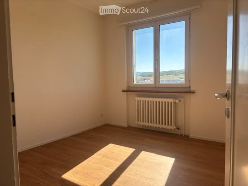 Empty room with wooden flooring, white walls, a window with a view, and a radiator under the window.