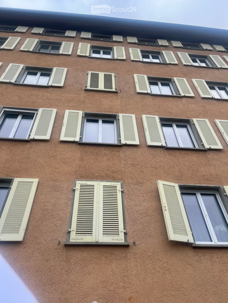 A section of a building facade with multiple windows, some with shutters open and others closed, featuring a textured brown exterior wall.