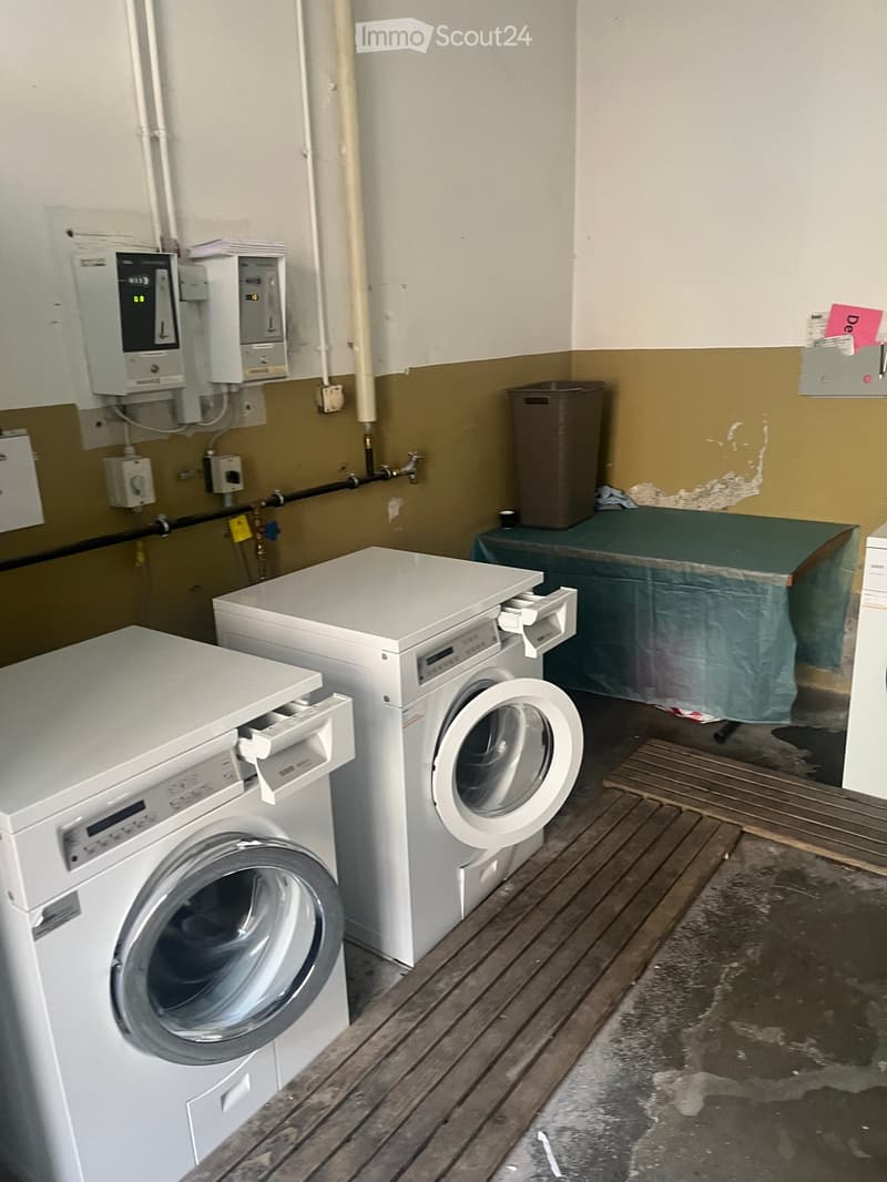 A small utility room with a washing machine, dishwasher, and a table covered with a green cloth. The floor is wooden, and there is a trash bin in the corner.