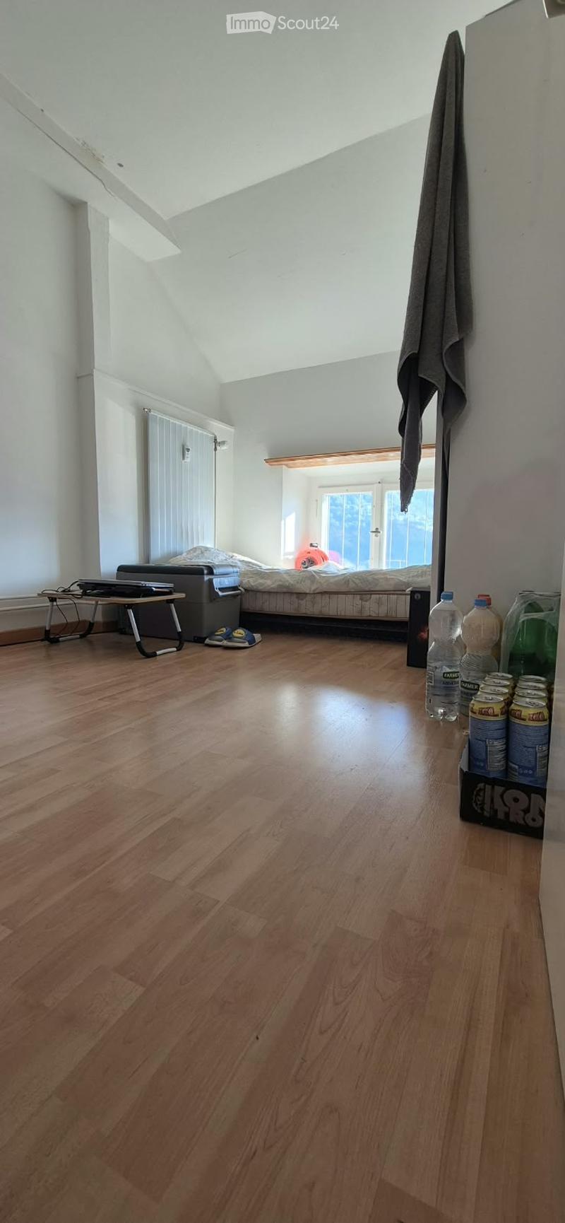 A furnished room with wooden flooring, a bed, a small table with a laptop, a radiator, and various items including bottles and cans.