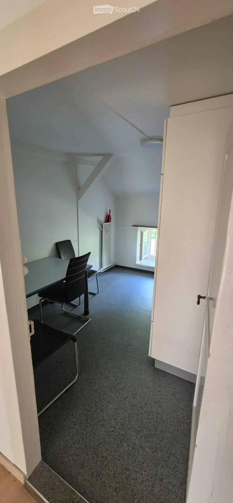 A small, furnished room with a slanted ceiling, featuring a table and chairs, a radiator, and a window letting in natural light.