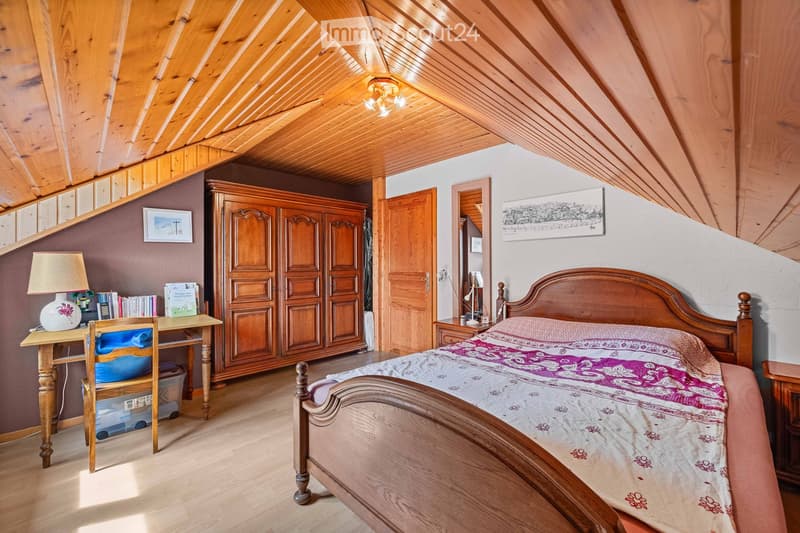 Cozy attic bedroom with wooden slanted ceilings, a large wooden bed, a wardrobe, a desk with a lamp, and a chair.