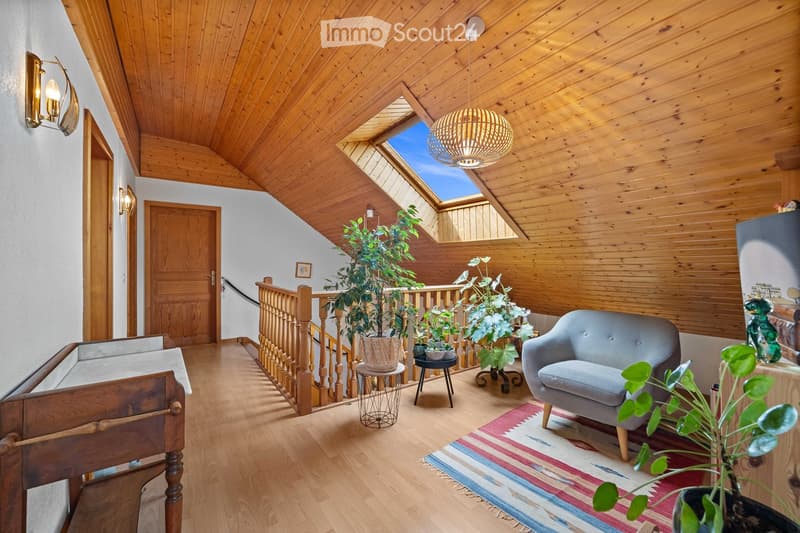 Cozy, wooden-paneled room with a slanted ceiling, skylight, and a staircase leading to an upper level. Features a grey sofa, small round table, potted plants, and a colorful rug.