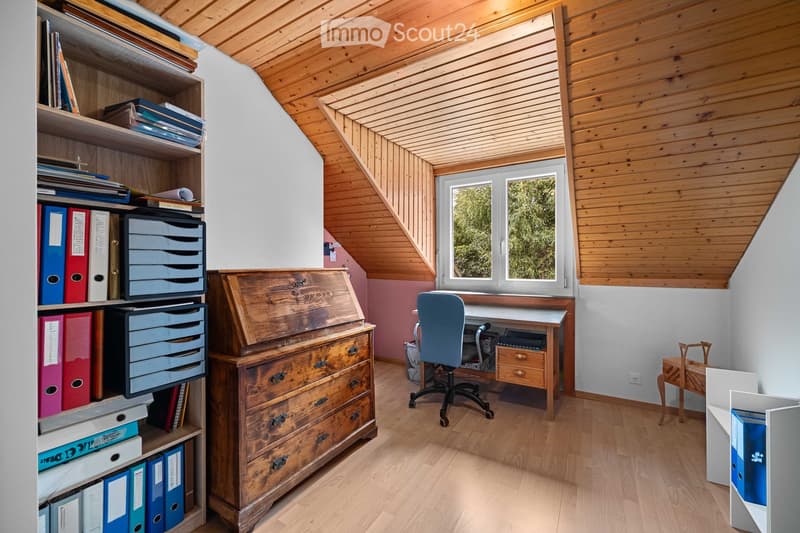 A cozy attic study with wooden beams, a large window, a wooden desk, a blue office chair, a bookshelf filled with books and folders, and a small wooden cabinet.