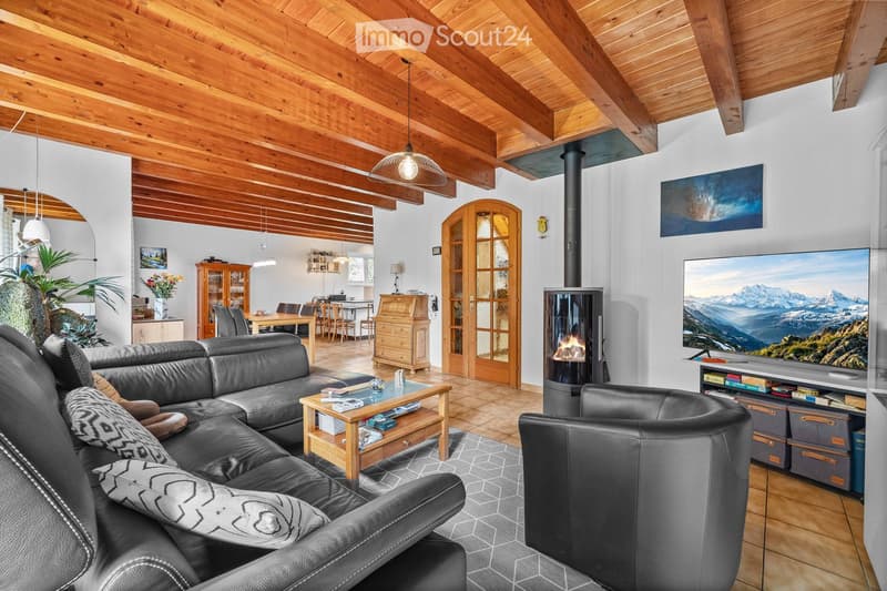 Living room with exposed wooden beams, a black leather sofa, a coffee table, an armchair, a fireplace, a large TV, and a dining area with a table and chairs.