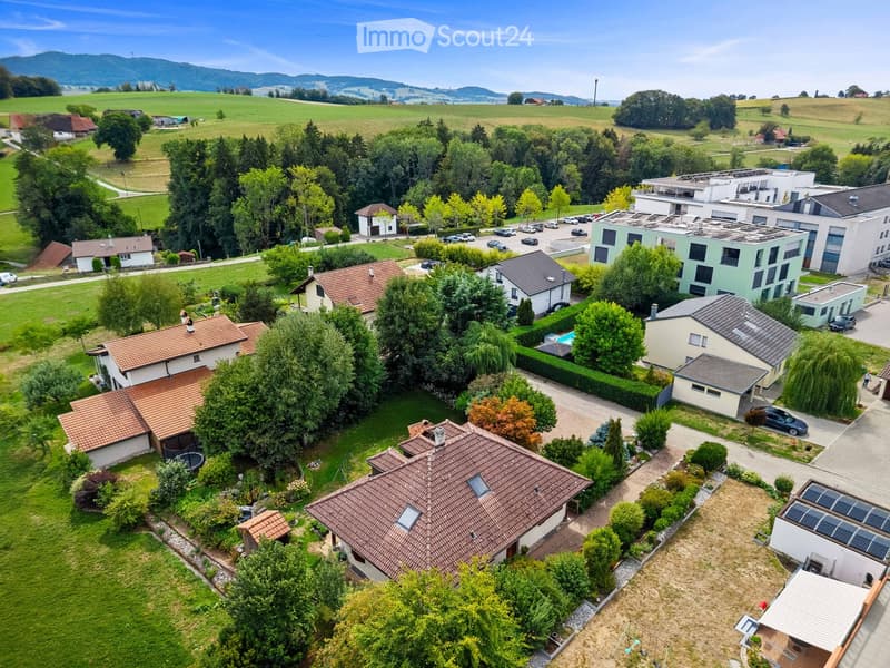 Aerial view of a residential area with multiple houses, gardens, and a swimming pool, surrounded by green fields and trees.