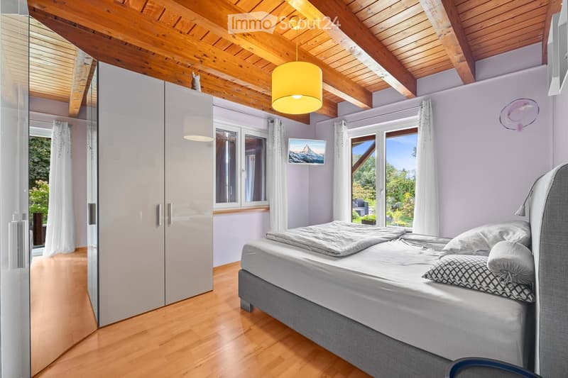 Bright bedroom with wooden floors, large windows, a grey bed with pillows and a duvet, a white wardrobe, and a hanging yellow lamp.