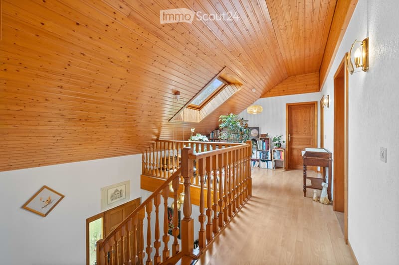 Spacious hallway with wooden staircase, wooden railings, and a skylight above. Features include a small table, bookshelves, and decorative wall art.