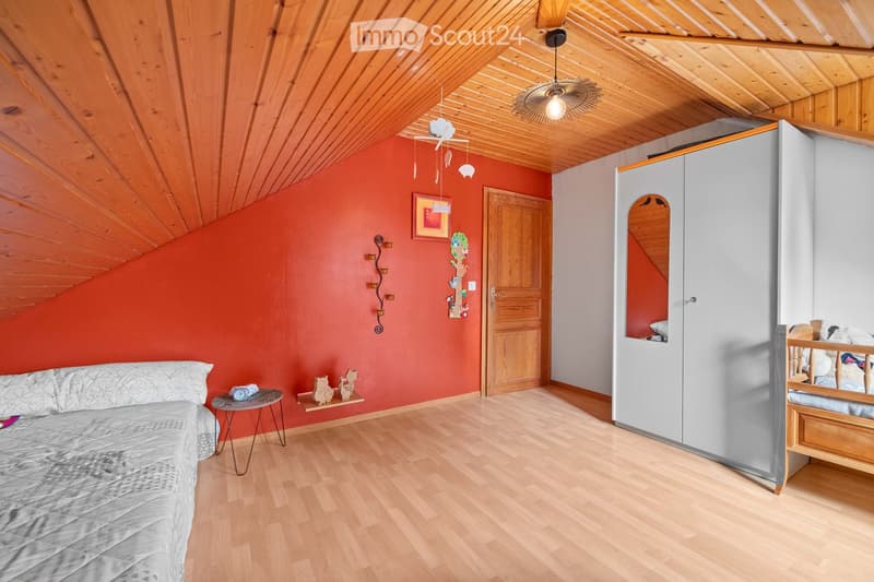 Bright bedroom with wooden floors, slanted wooden ceiling, red accent wall, white wardrobe with mirror, small table, and baby crib.