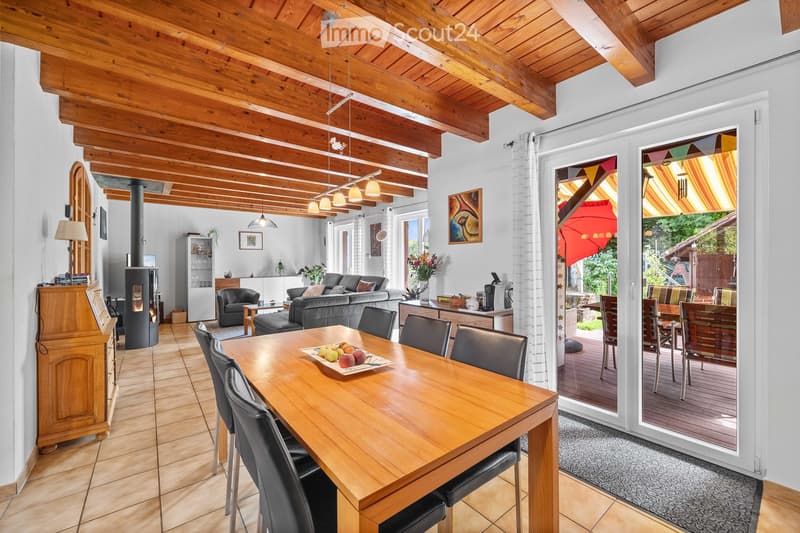 Bright living and dining area with exposed wooden beams, a large dining table with chairs, a sofa, an armchair, a fireplace, and a view to a patio.