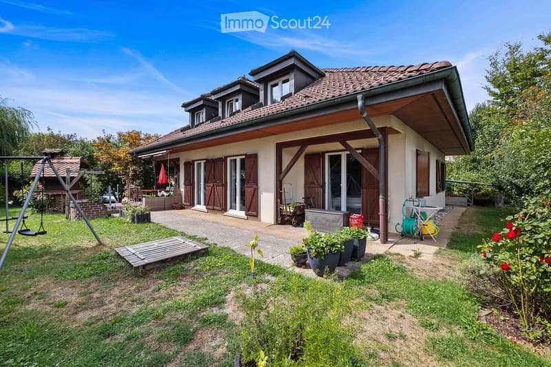 Two-story house with a tiled roof, wooden shutters, and a covered entrance. The garden includes a swing, potted plants, and a small patio area.