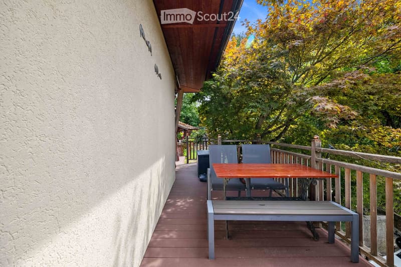 Balcony with wooden decking, a small table, two chairs, and a bench, surrounded by trees and greenery.