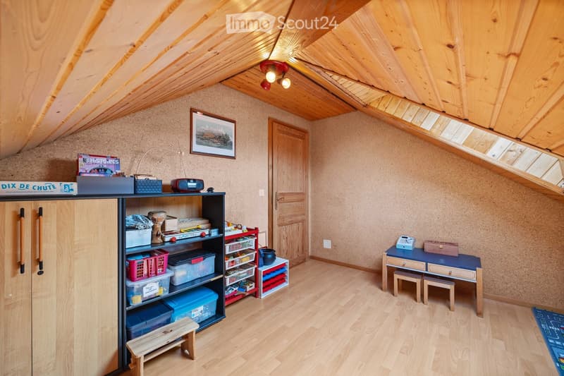 A cozy attic room with wooden floors, slanted wooden ceilings, and skylights. It features a cabinet, shelves filled with toys and storage bins, a small table with stools, and a radiator.