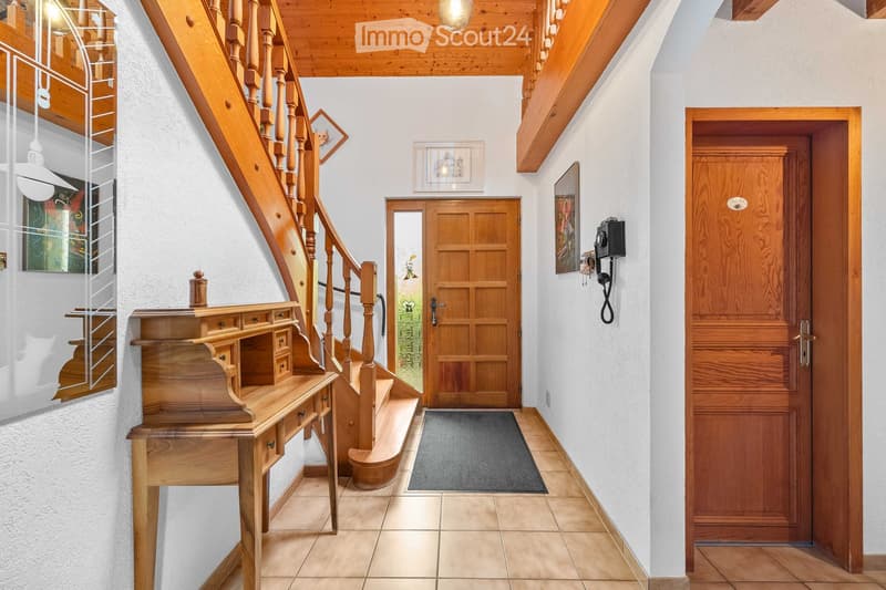 Spacious hallway with wooden staircase, wooden doors, and a small wooden desk. Light-colored walls and tiled flooring.