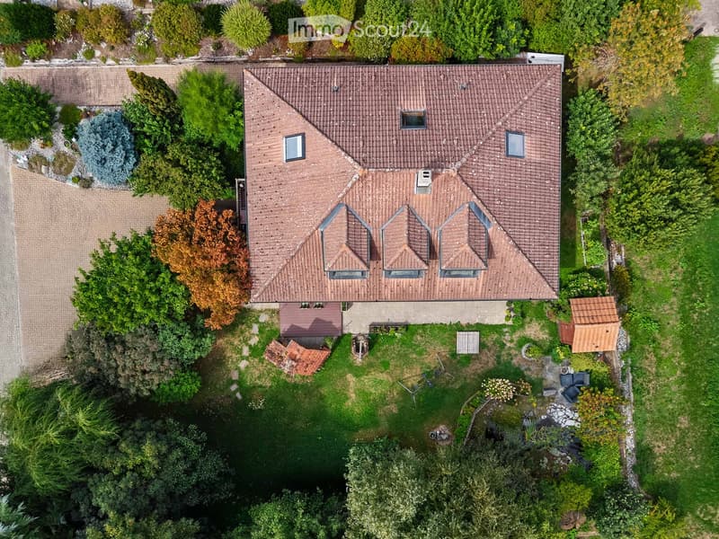 A two-story house with a red-tiled roof, surrounded by a well-maintained garden with trees, shrubs, and a small pond.