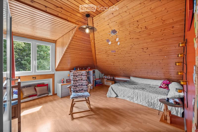 Cozy attic bedroom with wooden walls and ceiling, large windows, a bed with a quilt, a wooden chair, a small table, and a bookshelf.
