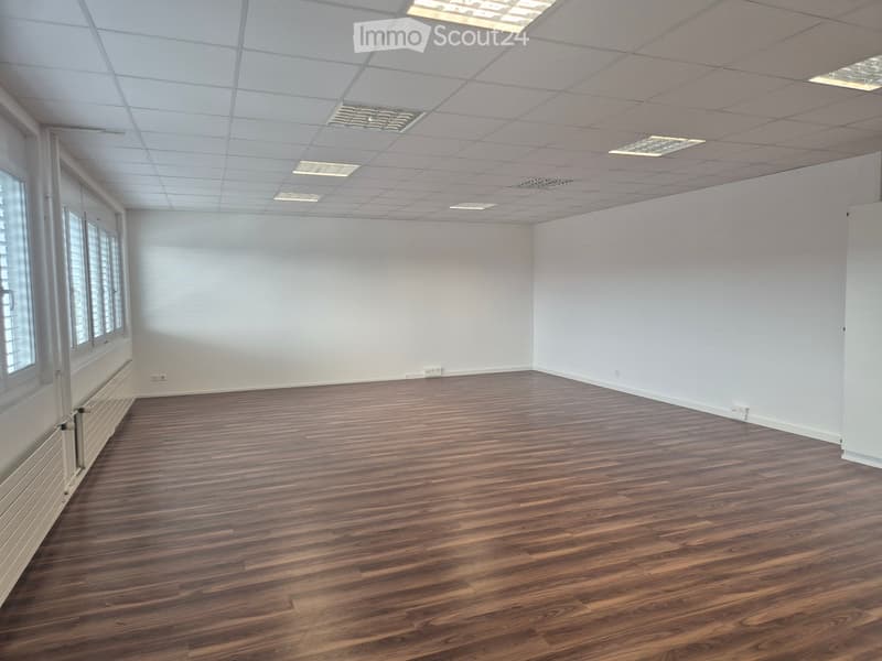 Empty room with wooden flooring, white walls, large windows, and a drop ceiling with recessed lighting.