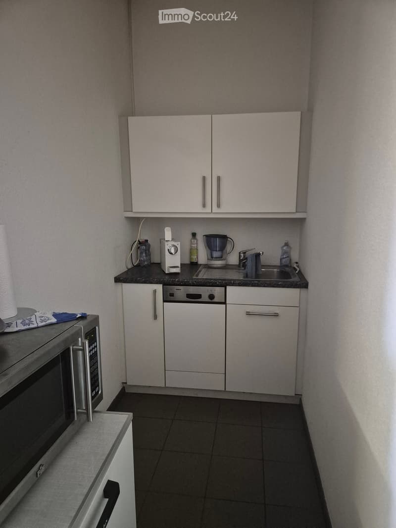 Compact kitchen with white cabinets, black countertop, microwave, dishwasher, sink, coffee maker, and various small appliances.