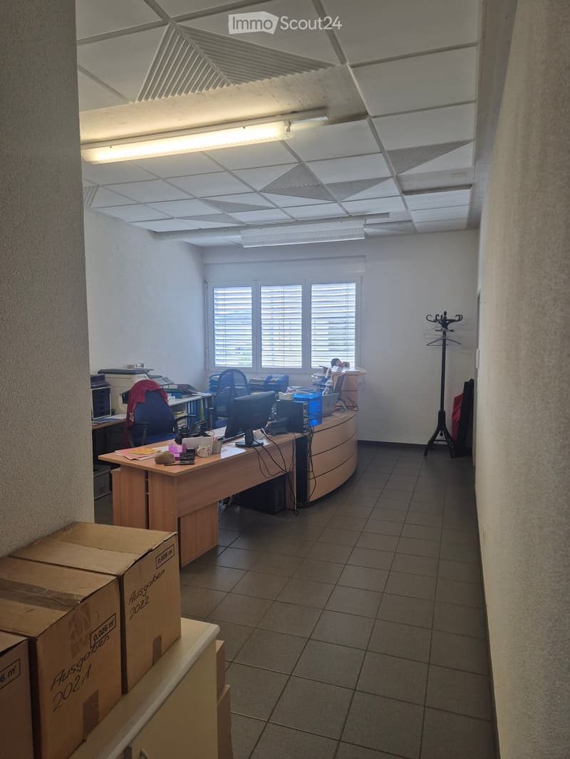 Office space with a curved wooden desk, multiple chairs, computers, and a coat rack. The room has tiled flooring and large windows with blinds.