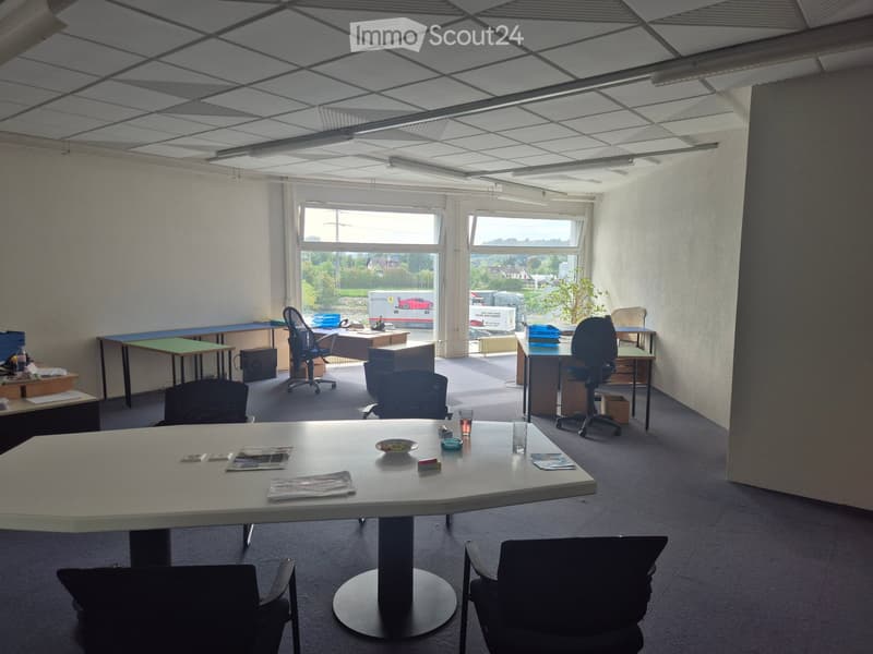 Office space with multiple tables, chairs, and office supplies, large windows with a view of the outside, and a modern ceiling with integrated lighting.
