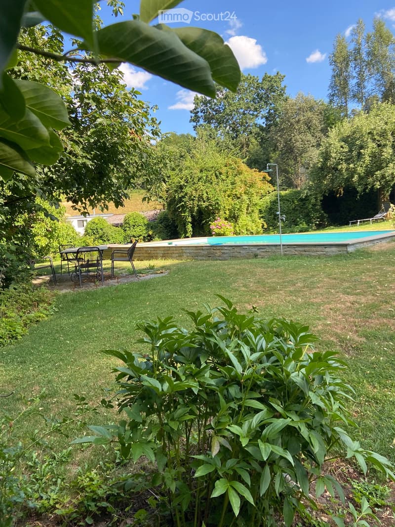 Well-maintained garden with a swimming pool, surrounded by lush greenery and trees. A small seating area with chairs and a table is visible near the pool.