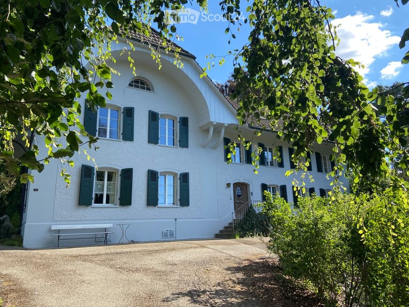 A two-story white house with green shutters, large windows, and a curved roofline. The house features a front bench and a small table near the entrance, surrounded by greenery and trees.