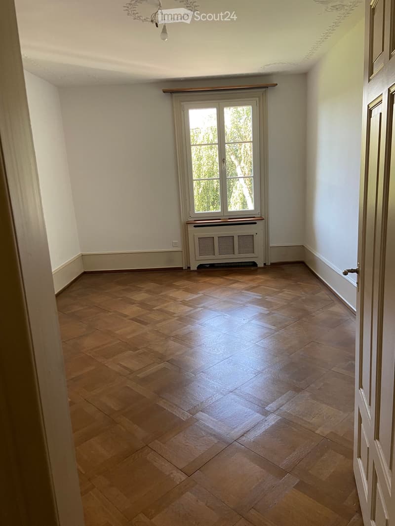 Empty room with wooden parquet flooring, white walls, and a large window with double doors leading outside. A radiator is visible beneath the window, and a chandelier hangs from the ceiling.