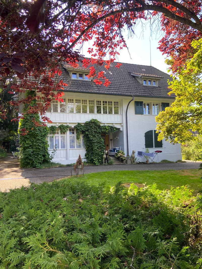 Two-story house with white exterior, multiple windows, and a well-maintained garden with various plants and trees.
