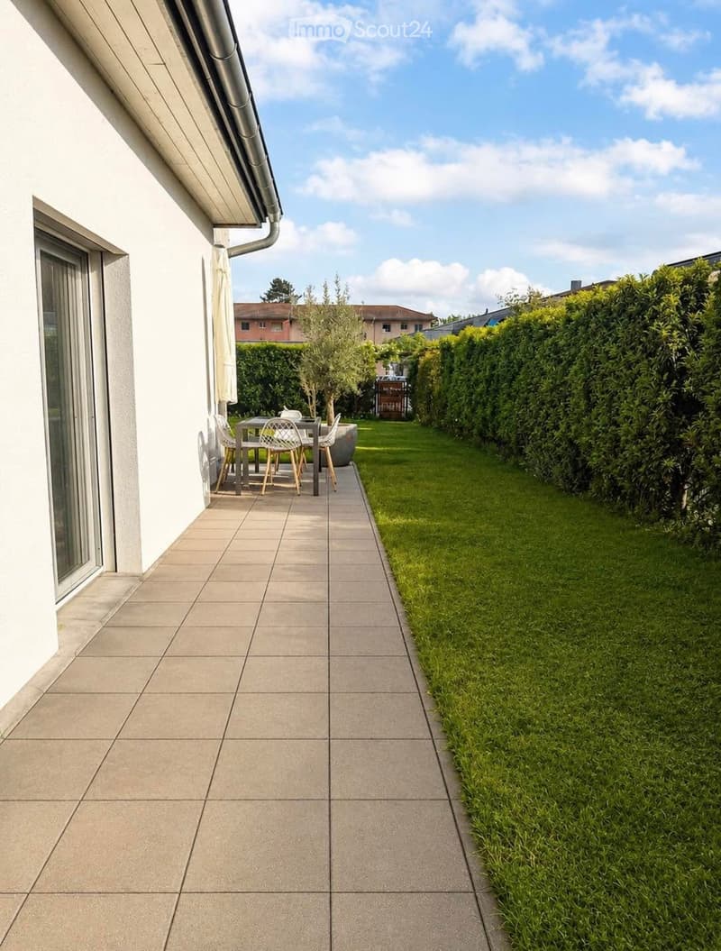 Outdoor patio area with tiled flooring, a small table and chairs, surrounded by a well-maintained lawn and hedges.