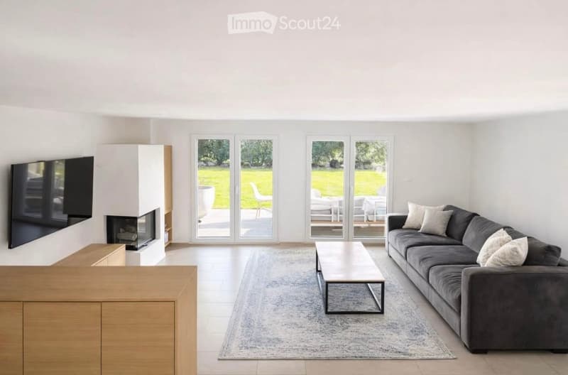 Modern living room with large windows offering a view of a green outdoor area, featuring a grey sectional sofa, a wooden coffee table, a fireplace, and a mounted TV.