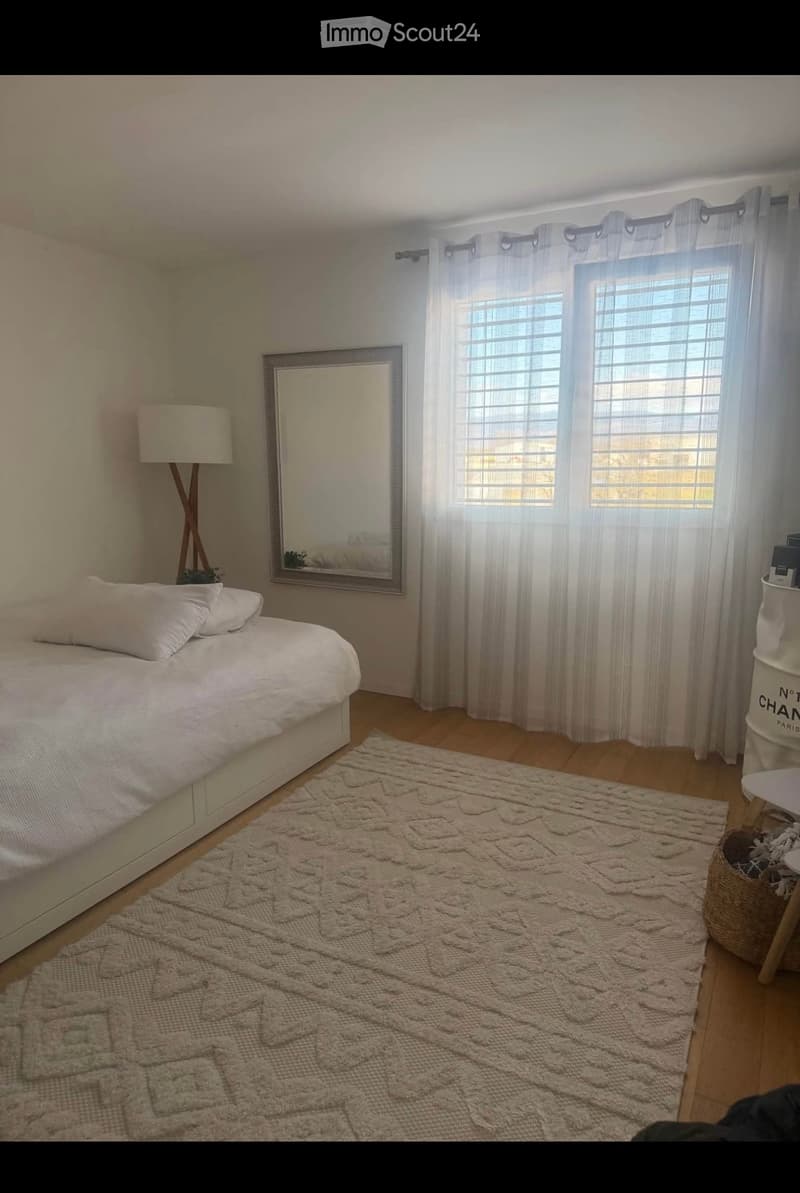 Cozy bedroom with a neatly made bed, a large mirror, a floor lamp, a patterned rug, and a small stool with a basket on it.