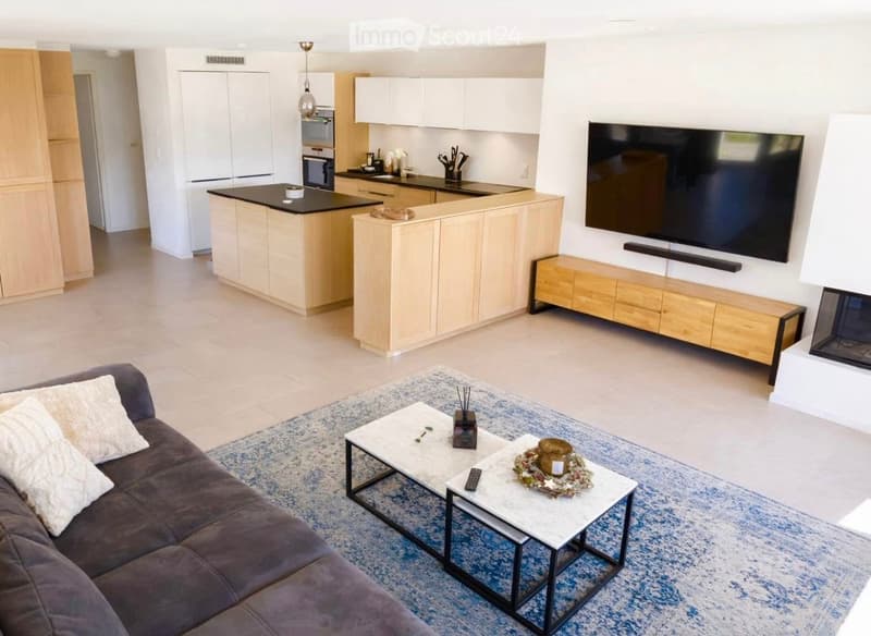 Open-plan living and dining area with a sofa, coffee table, and a large television. A modern kitchen with white cabinets and a black countertop is visible in the background, along with a fireplace.