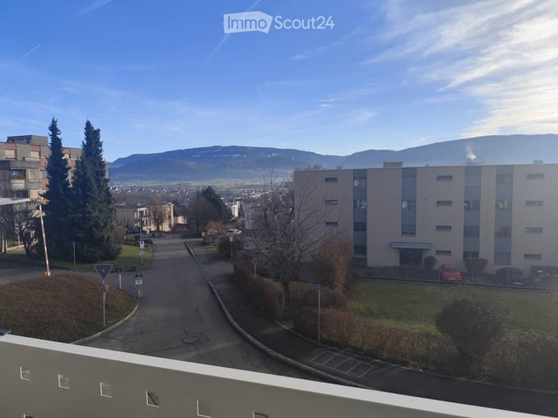 Aerial view of a building with a balcony, surrounded by trees, mountains, and a road with parked cars.