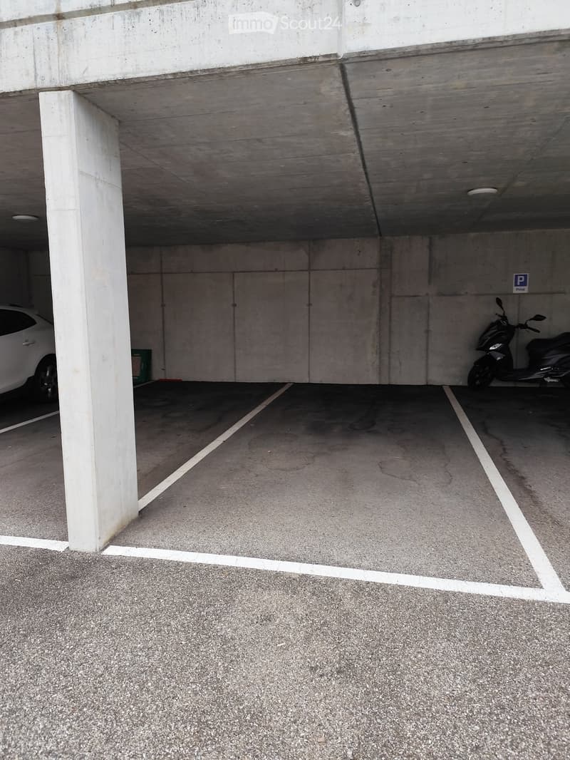 An underground parking lot with a concrete ceiling, white columns, and marked parking spaces. A white car and a scooter are parked inside the lot.